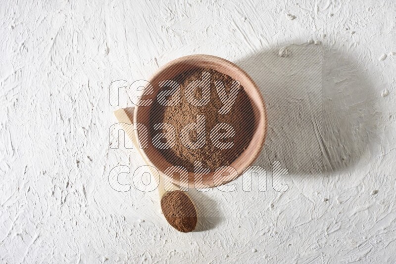 A wooden bowl and a wooden spoon full of cloves powder on a textured white flooring