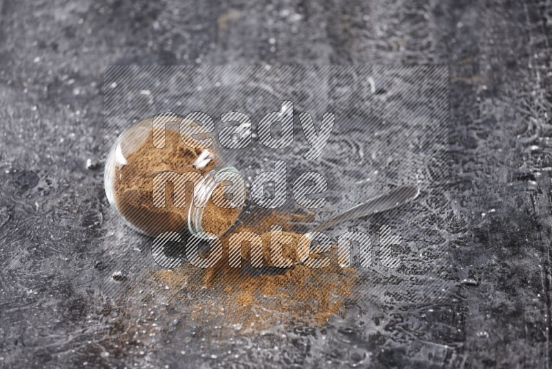 Herbal glass jar full of cinnamon powder flipped and a metal spoon on textured black background