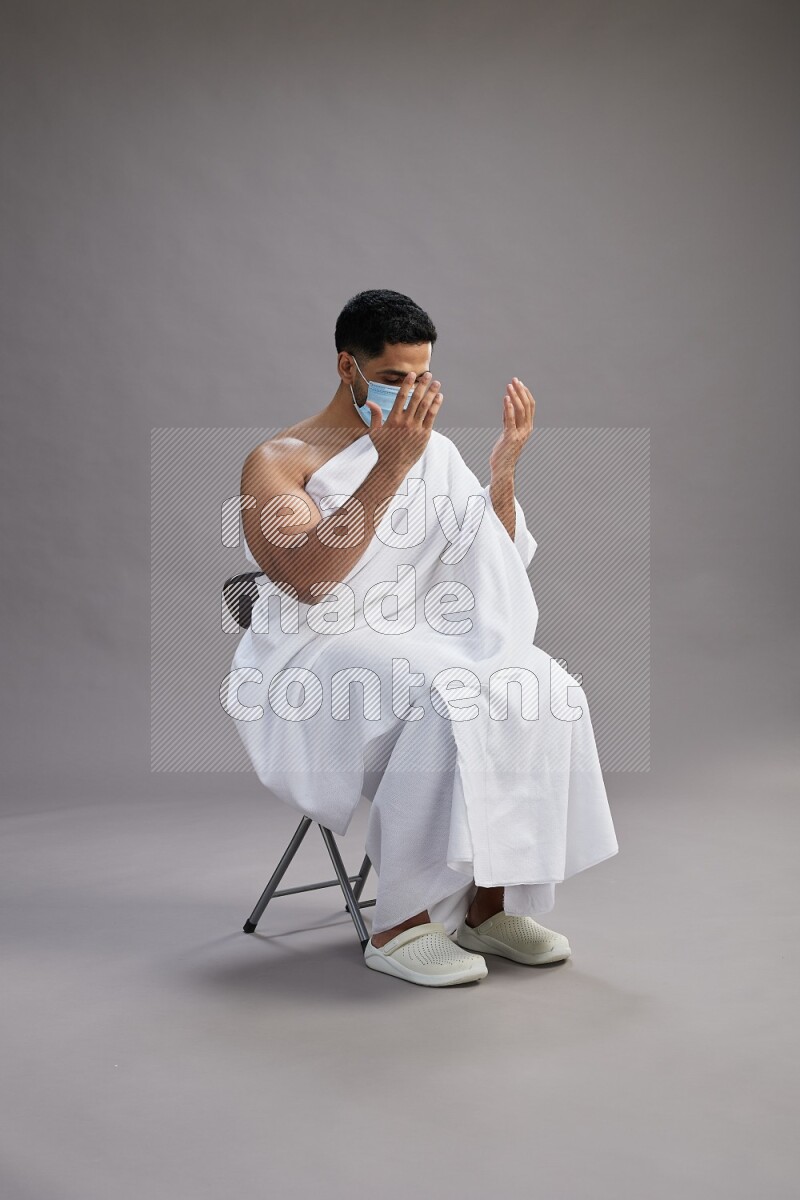 A man wearing Ehram with face mask sitting on chair dua'a on gray background