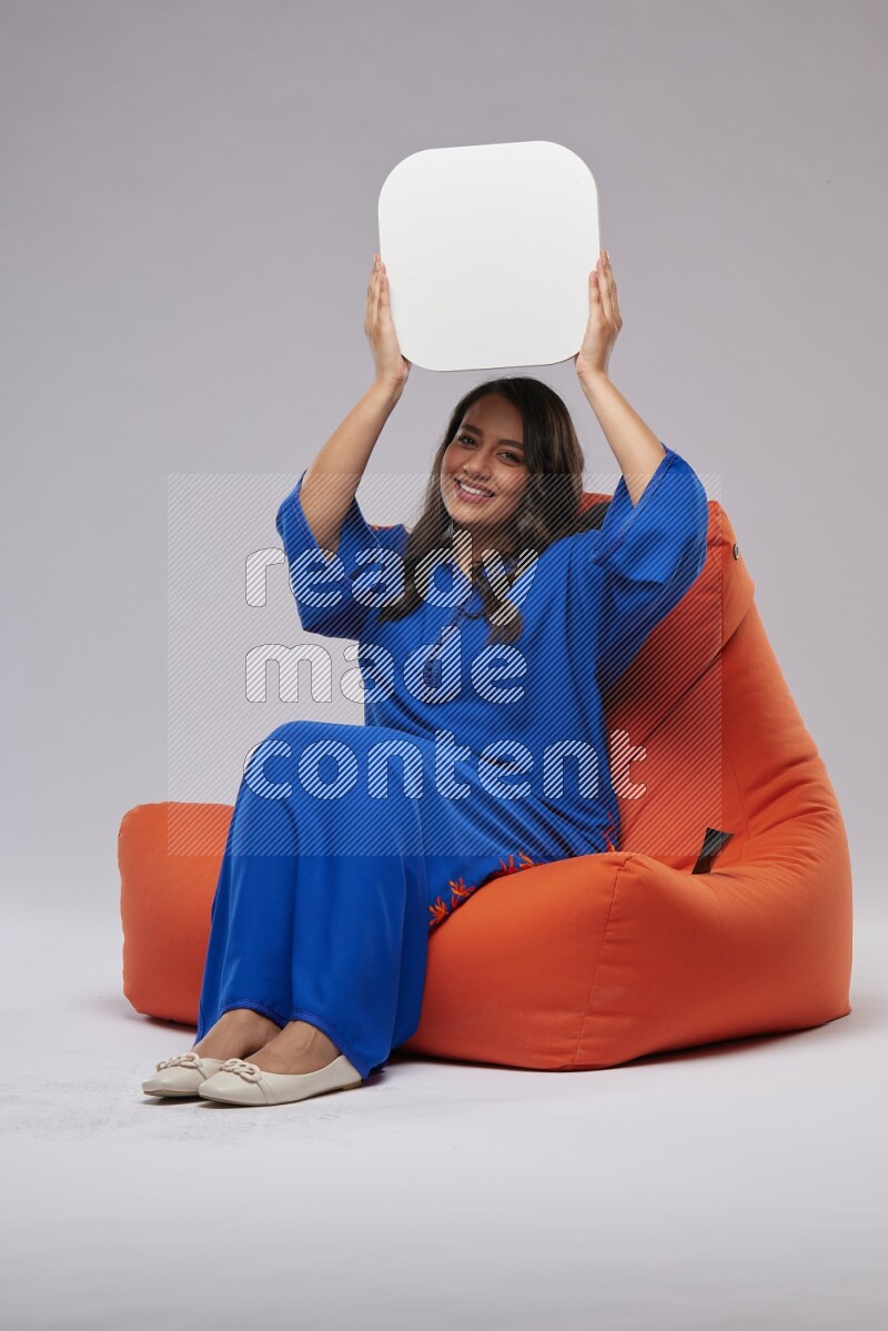 A Woman sitting on an orange beanbag wearing Jalabeya holding a social media sign