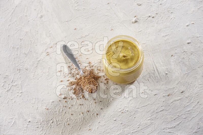 A glass jar full of mustard paste and a metal spoon full of mustard seeds on a textured white flooring