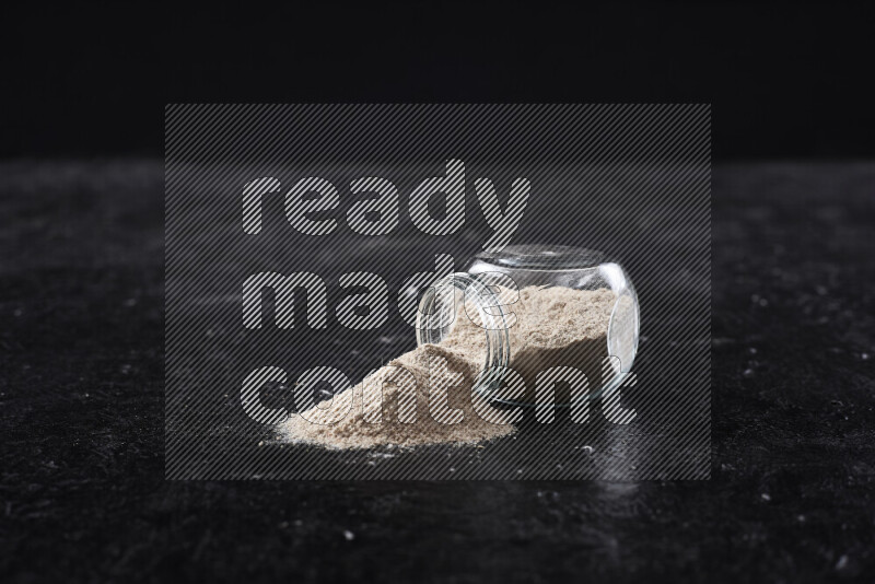 A glass jar full of onion powder flipped with some spilling powder on black background
