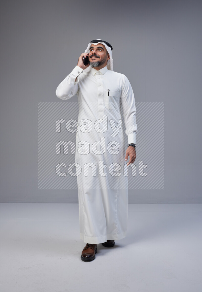 Saudi man Wearing Thob and white Shomag standing talking on phone on Gray background