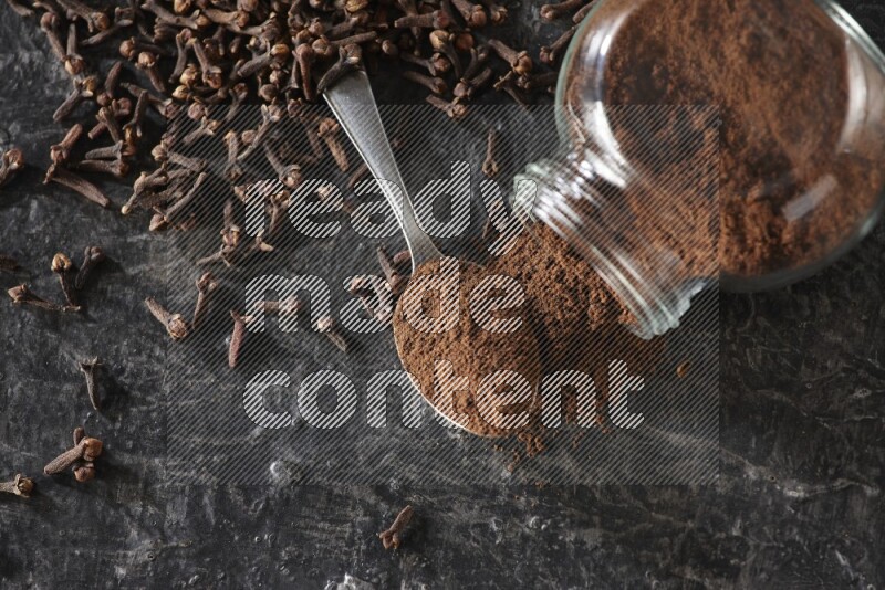 A flipped glass spice jar and a metal spoon full of cloves powder with cloves spread on textured black flooring