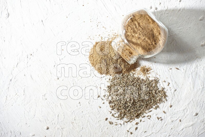 A flipped glass spice jar full of cumin powder and powder spilled out with cumin seeds on textured white flooring