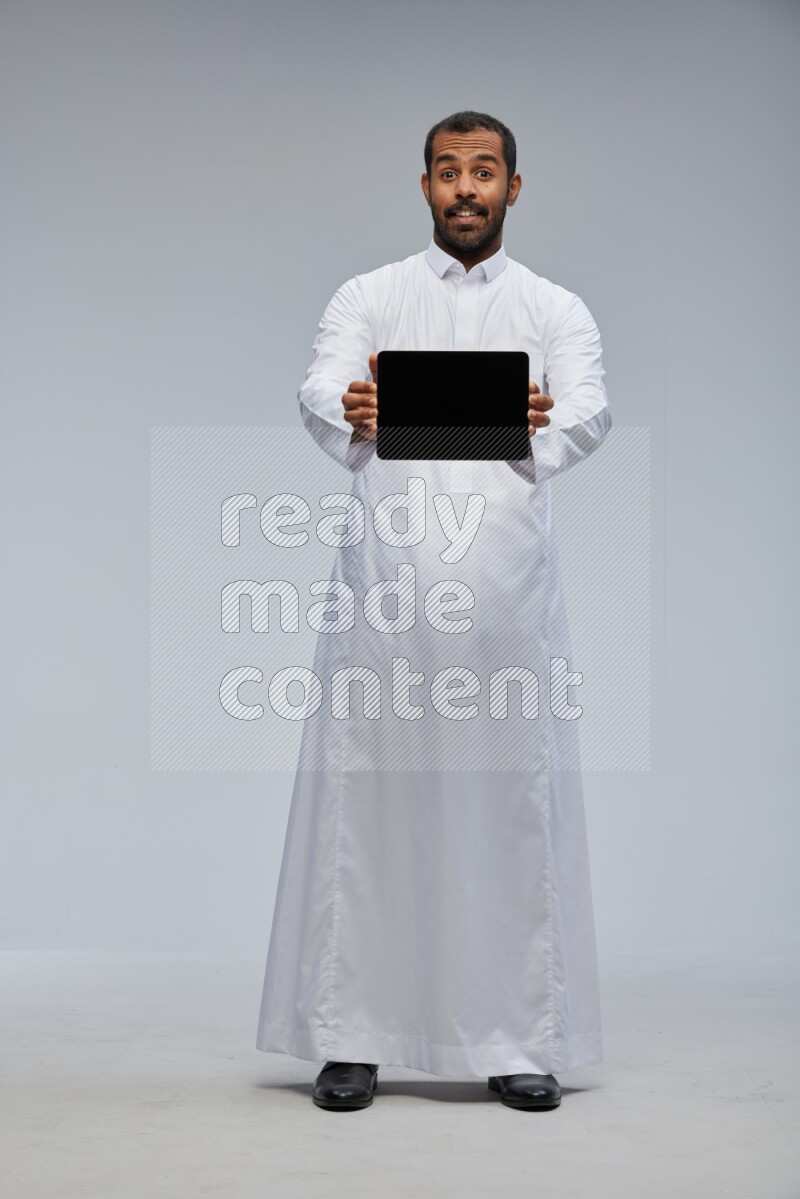 Saudi man wearing Thob standing showing tablet to camera on Gray background