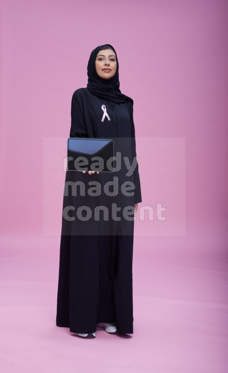 Saudi woman wearing pink ribbon on Abaya standing showing a tablet to camera on pink background