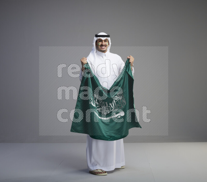 A Saudi man standing wearing thob and white shomag with face painting holding big Saudi flag on gray background