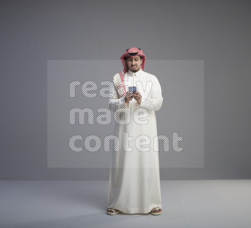 A  Saudi man standing wearing thob and red shomag texting on phone on gray background
