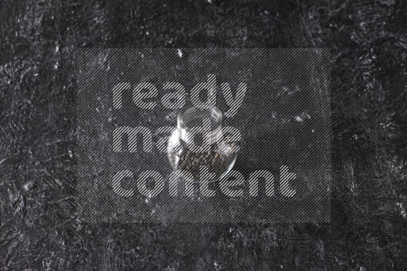 A glass spice jar full of black pepper beads on textured black flooring