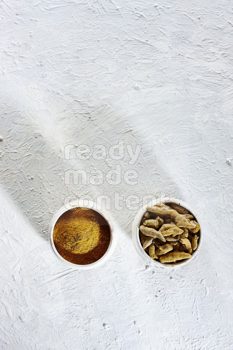 وعاءان فخاريان بيج ممتلئان ببودرة الكركم وأصابع الكركم المجففة على خلفية بيضاء