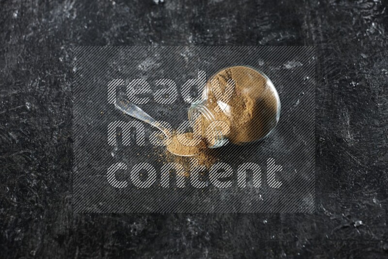 وعاء زجاجي للتوابل مقلوب وملعقة معدنية ممتلئة ببودرة الكمون والبودرة تناثرت منه على خلفية سوداء