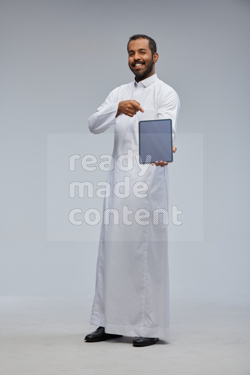 Saudi man wearing Thob standing showing tablet to camera on Gray background