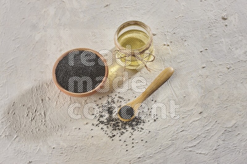 A wooden bowl and spoon full of black seeds with a glass jar of black seeds oil on a textured white flooring