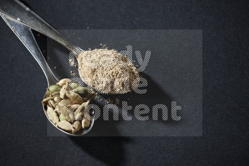 2 Metal spoons full of cardamom powder and cardamom seeds on black flooring
