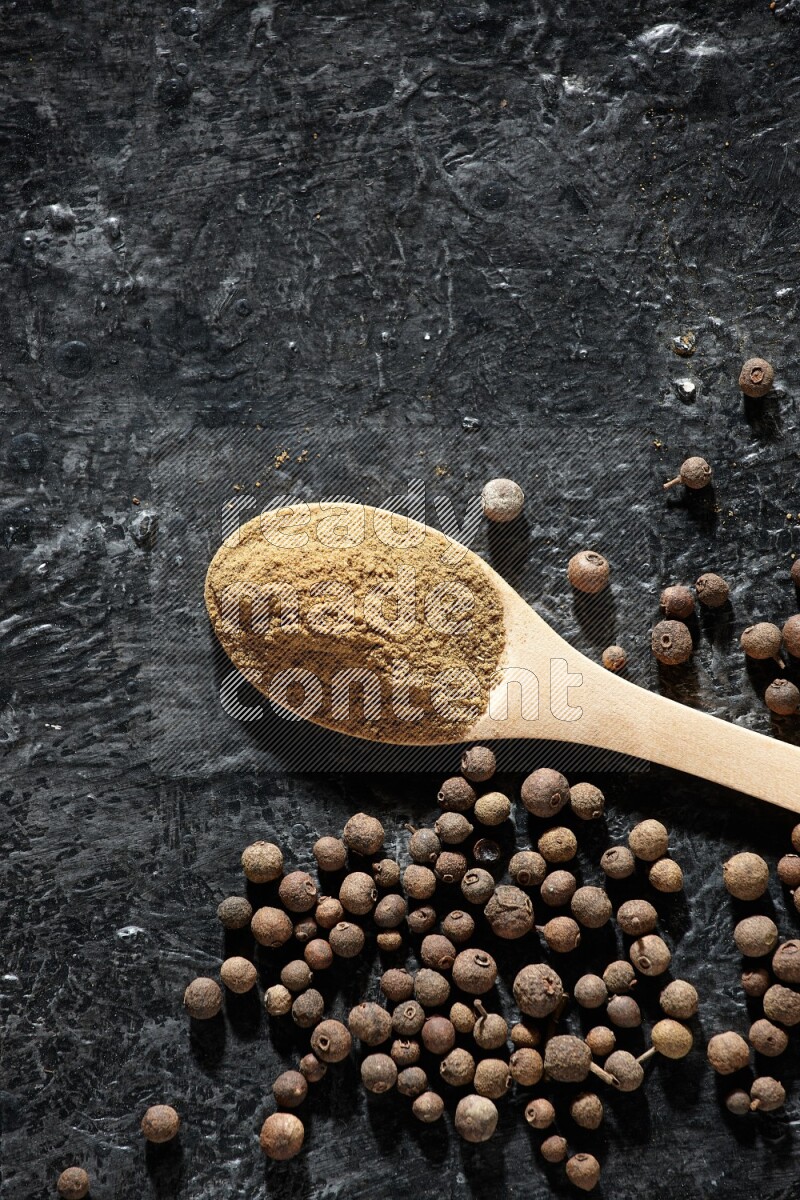 Wooden spoons full of all spice powder and allspice whole balls beside it on a textured black flooring