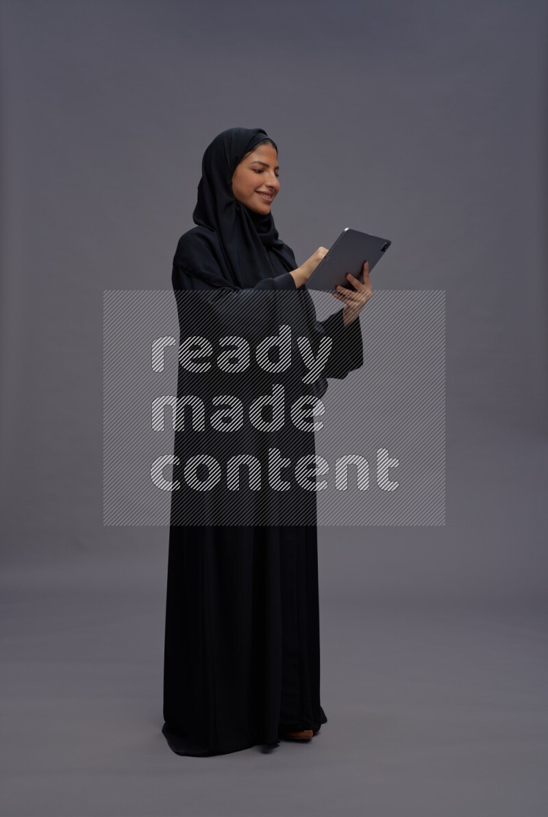 Saudi woman wearing Abaya standing working on tablet on gray background