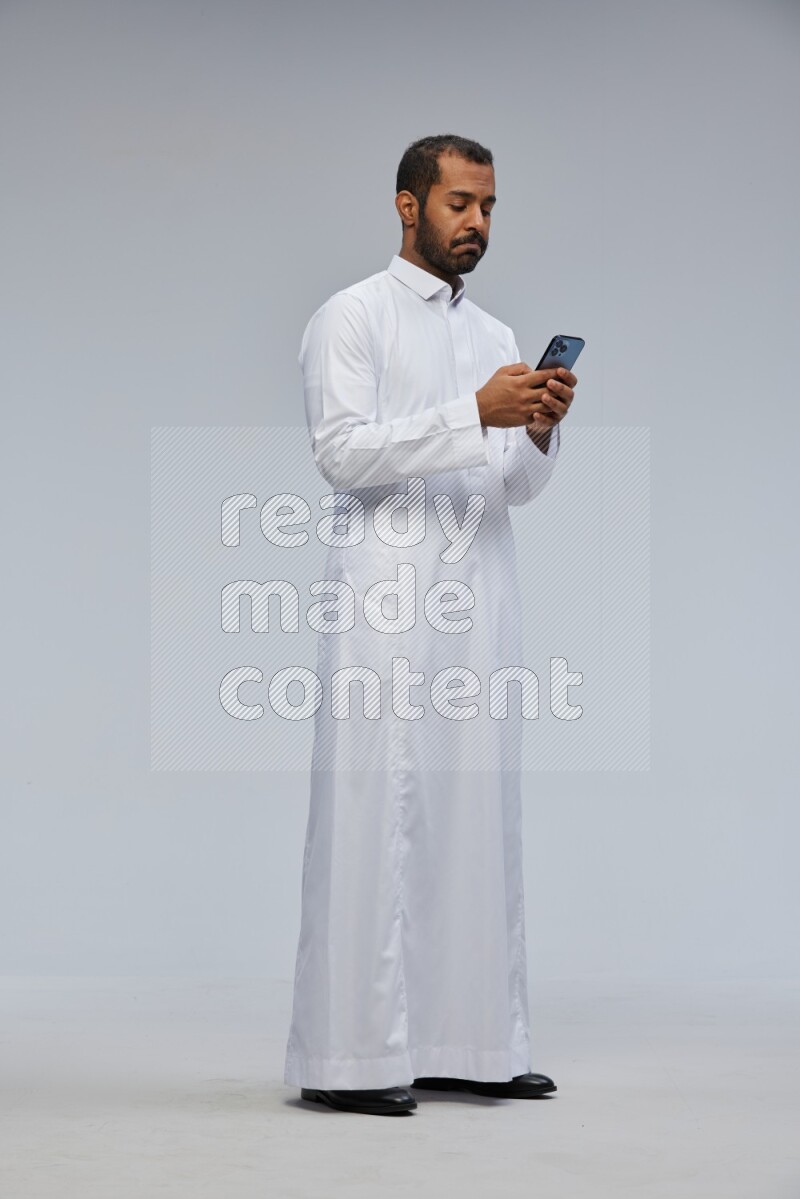 Saudi man wearing Thob standing texting on phone on Gray background
