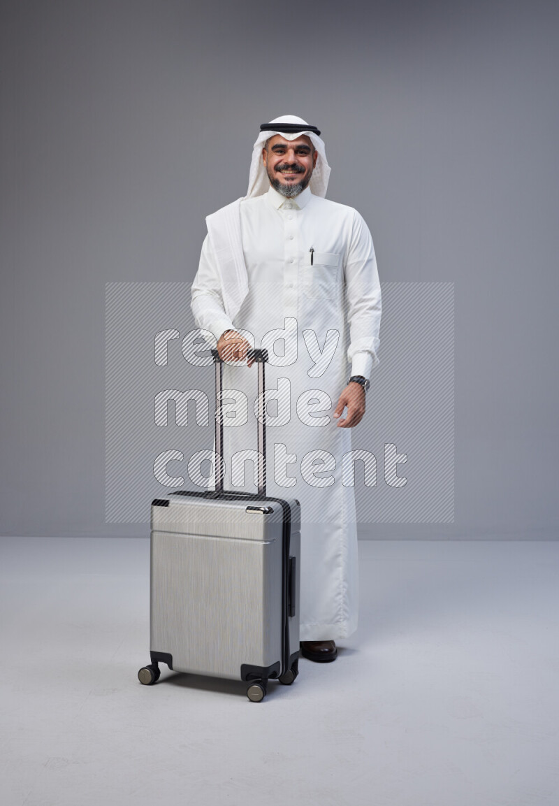 Saudi man wearing Thob and white Shomag standing holding Travel bag on Gray background