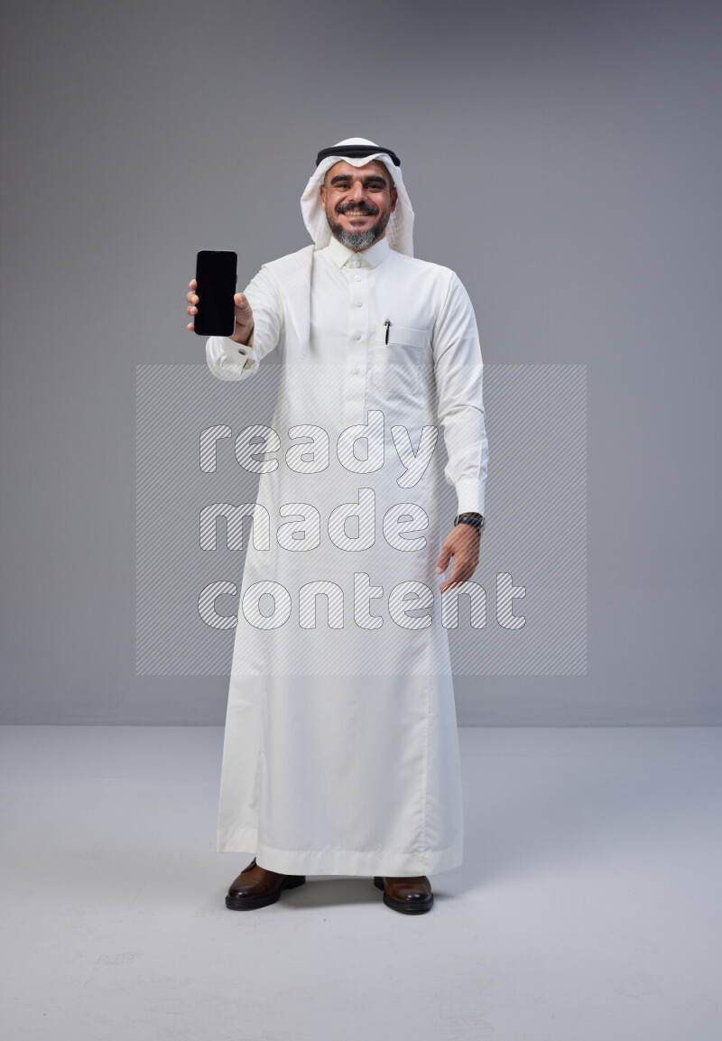 Saudi man Wearing Thob and white Shomag standing showing phone to camera on Gray background