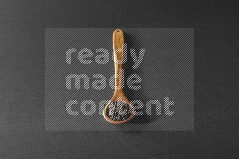 A wooden ladle full of black pepper beads on a black flooring