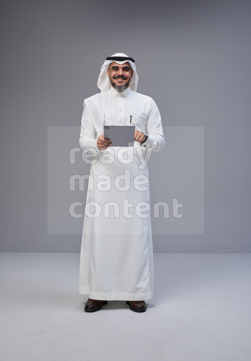 Saudi man Wearing Thob and white Shomag standing working on tablet on Gray background