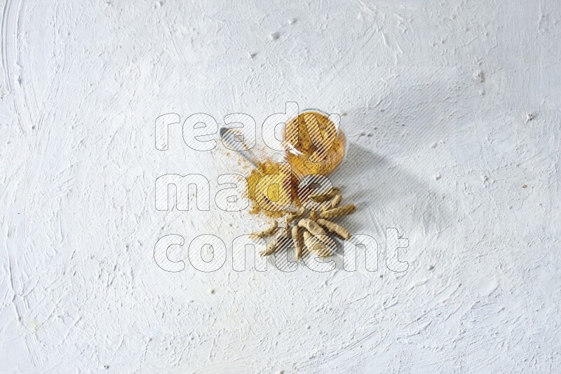 وعاء زجاجي للتوابل مقلوب وملعقة معدنية ممتلئة ببودرة الكركم والبودرة تناثرت منه مع أصابع الكركم المجففة على خلفية بيضاء