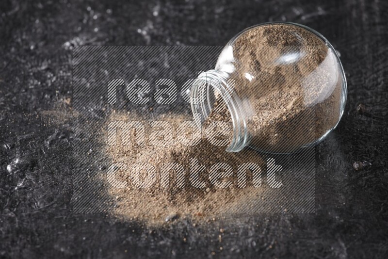 A flipped glass spice jar full of black pepper powder on textured black flooring