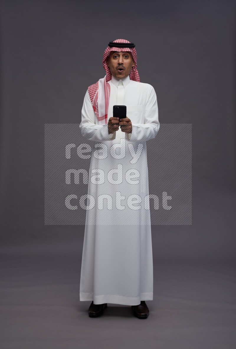 Saudi man wearing thob and shomag standing texting on phone on gray background