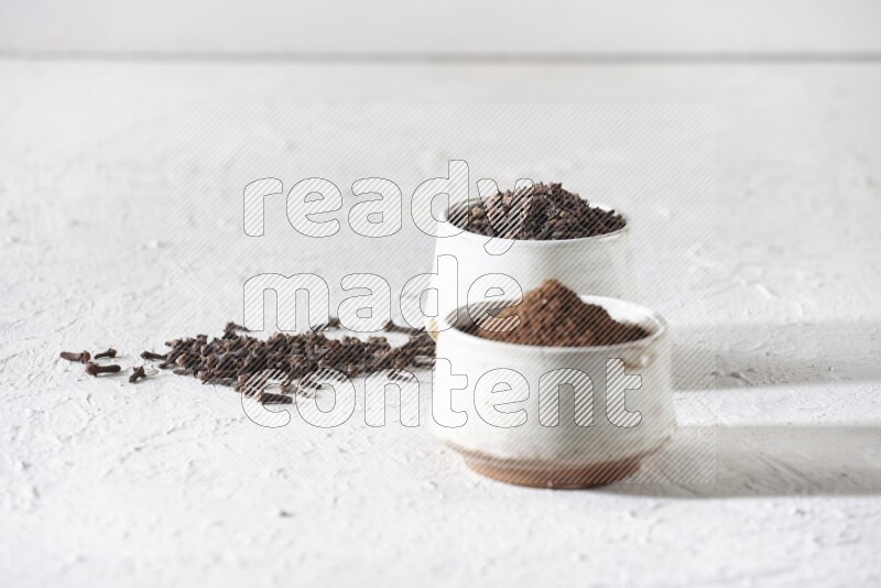 2 Beige ceramic bowls, one full of cloves and the other full of powder on textured white flooring