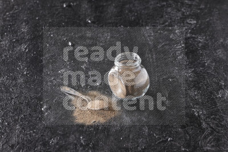 A glass spice jar full of black pepper powder and a metal spoon full of powder on textured black flooring