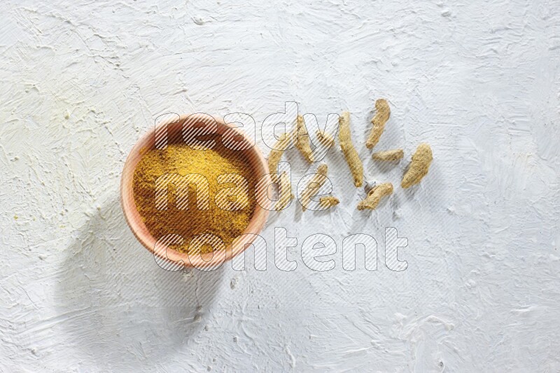 A wooden bowl full of turmeric powder with dried whole fingers on textured white flooring