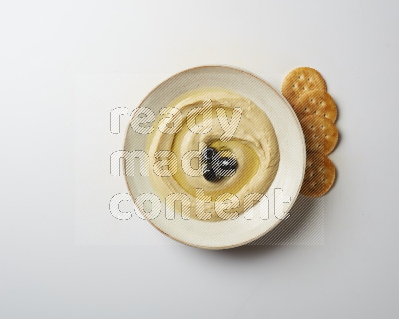 Hummus in a pottry plate garnished with black olives on a white background