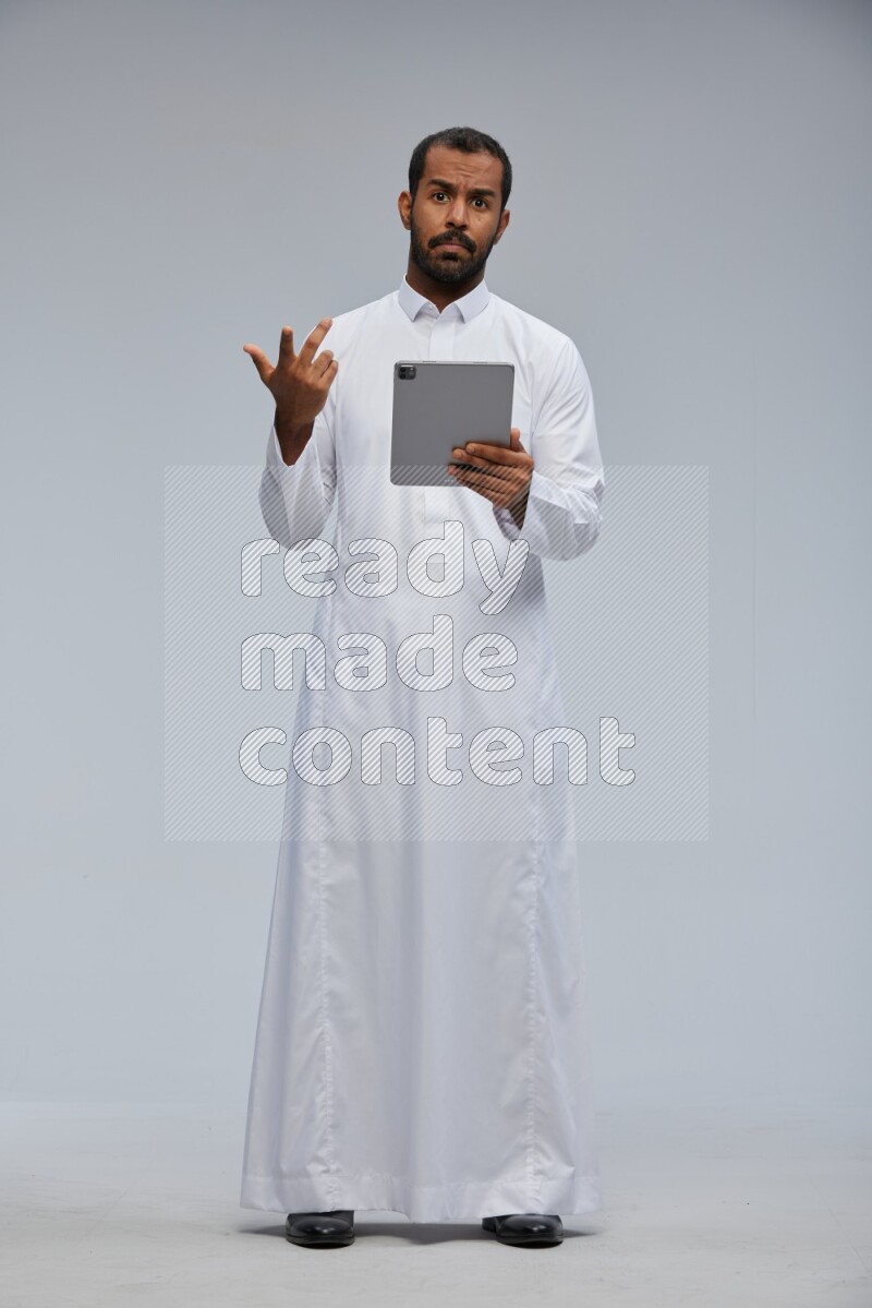 Saudi man wearing Thob standing working on tablet on Gray background