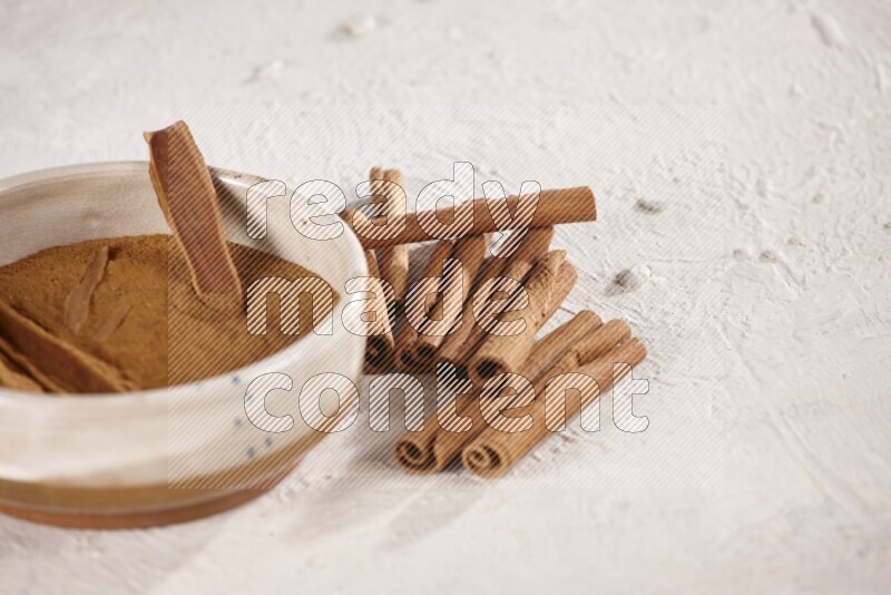 Ceramic bowl full of cinnamon powder with cinnamon sticks on the side on white background