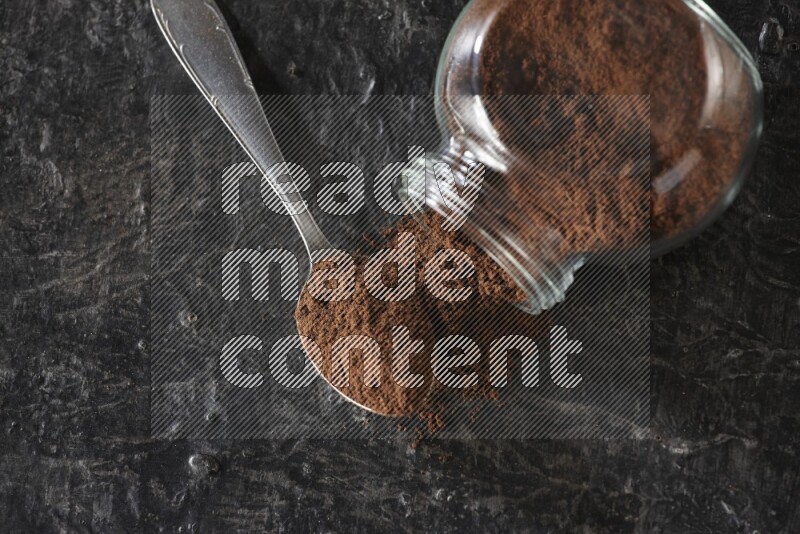 A flipped glass spice jar and a metal spoon full of cloves powder on textured black flooring
