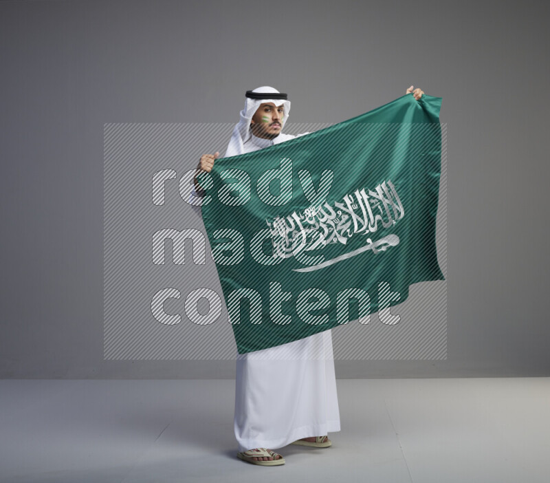A Saudi man standing wearing thob and white shomag with face painting holding big Saudi flag on gray background