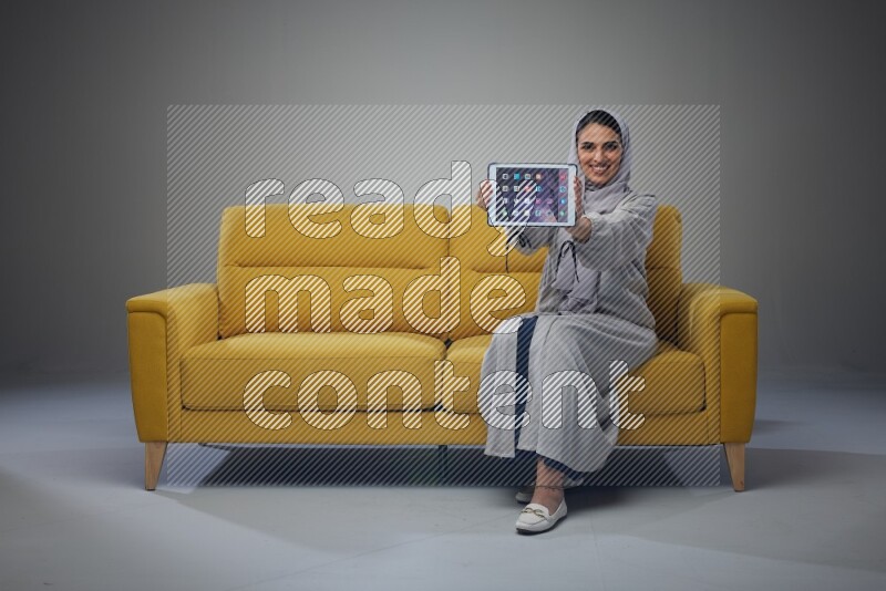 A Saudi female wearing a light gray Abaya and head scarf sitting on a yellow sofa and showing her tablet's screen while wearing headphones eye level on a grey background