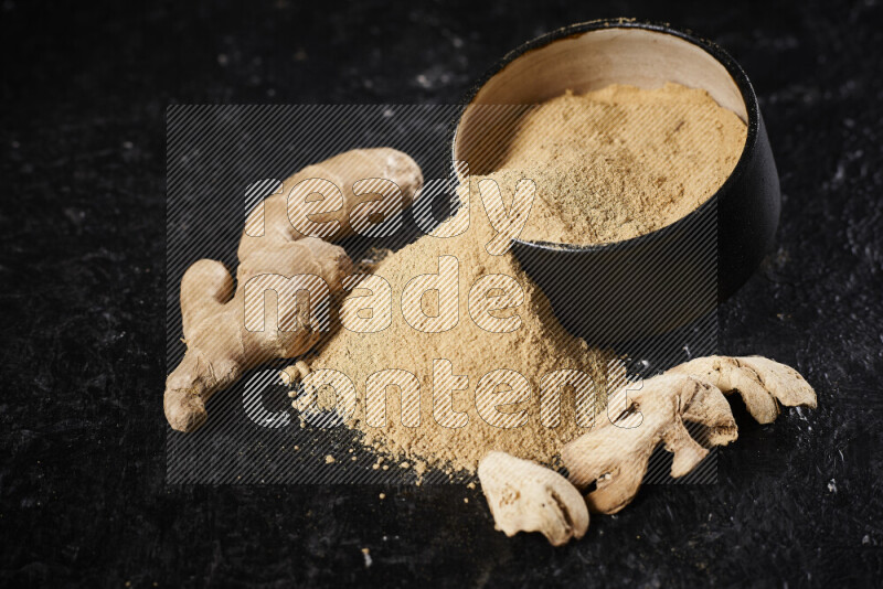 A black pottery bowl full of ground ginger powder with fallen powder from it on black background