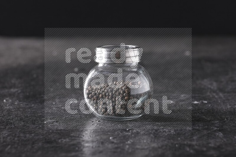 A glass spice jar full of black pepper beads on textured black flooring