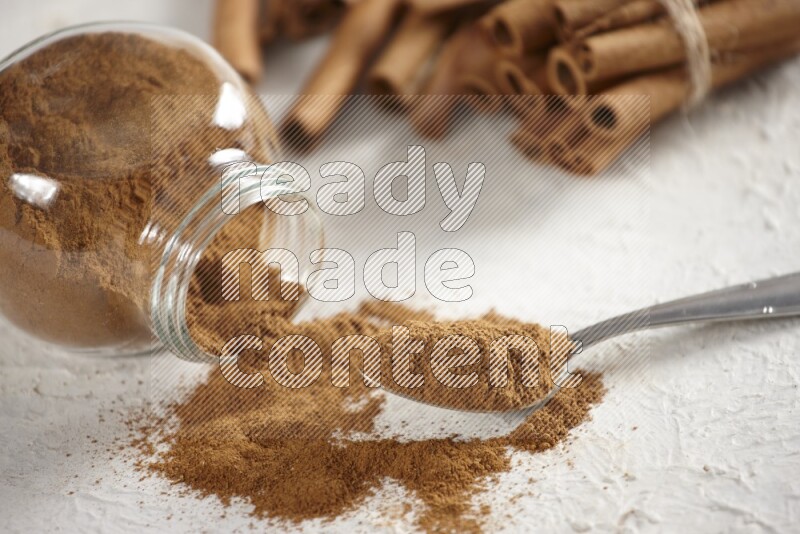 Flipped herbs glass jar full of cinnamon powder with a metal spoon full of powder and cinnamon sticks on a textured white background