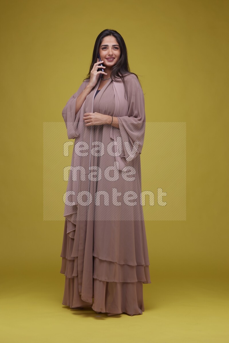 A woman Calling on a Yellow Background wearing Brown Abaya