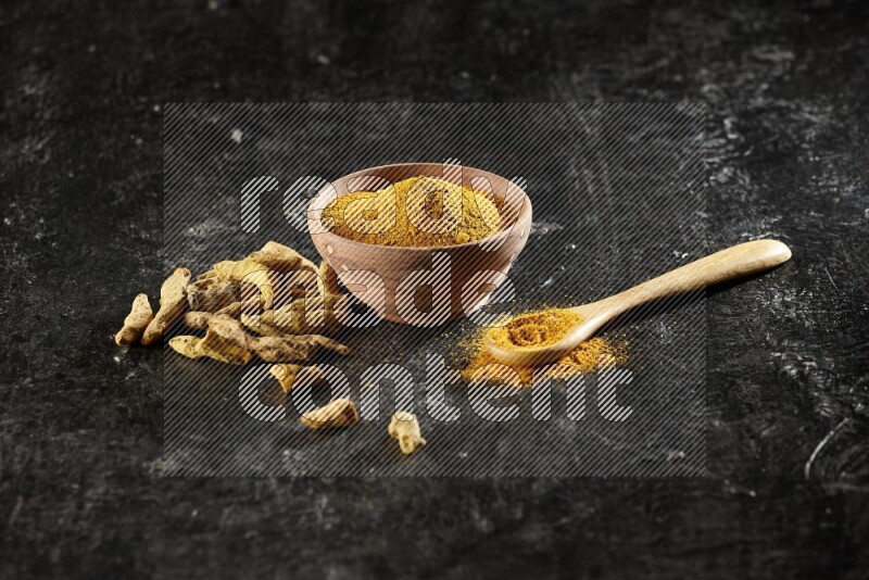 A wooden bowl and a wooden spoon full of turmeric powder with dried turmeric fingers on textured black flooring