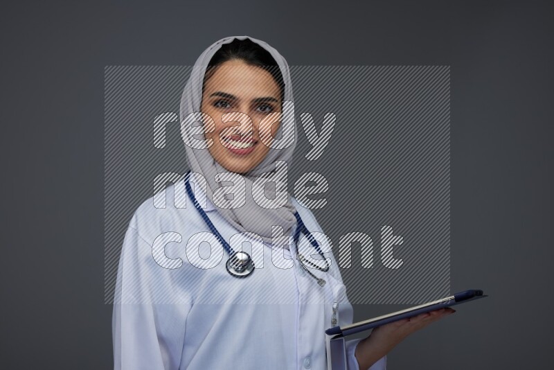 A Saudi doctor wearing a light gray head scarf standing and crossing her hands eye level on a grey background