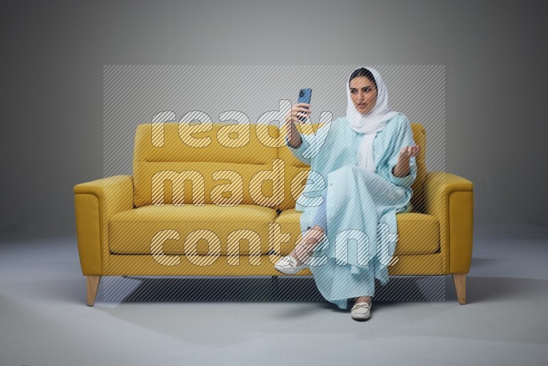 A Saudi woman wearing a light blue Abaya and white head scarf sitting on a yellow sofa and taking selfies with her phone eye level on a grey background