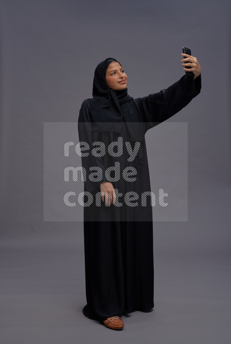 Saudi woman wearing Abaya standing taking selfie on gray background