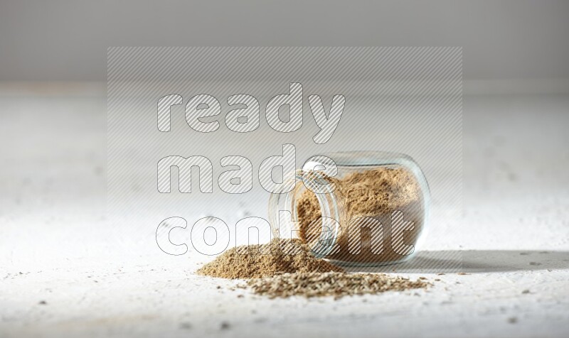 A flipped glass spice jar full of cumin powder and powder spilled out with cumin seeds on textured white flooring