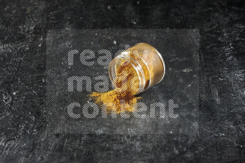 A flipped glass jar full of turmeric powder and powder spilled out of it on a textured black flooring