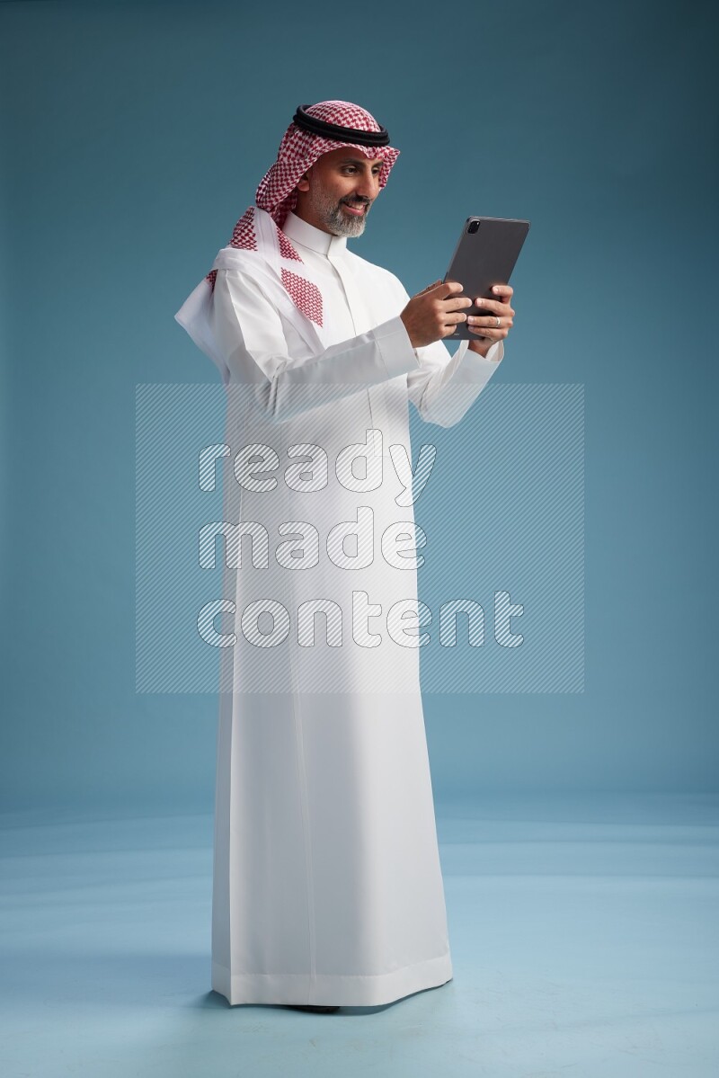 Saudi man wearing thob and shomagh with tablet on Blue background