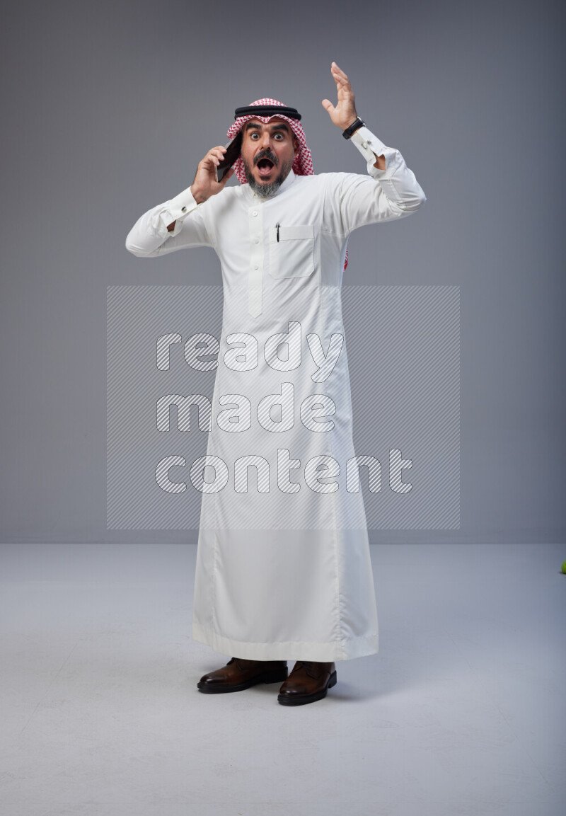Saudi man Wearing Thob and red Shomag standing talking on phone on Gray background